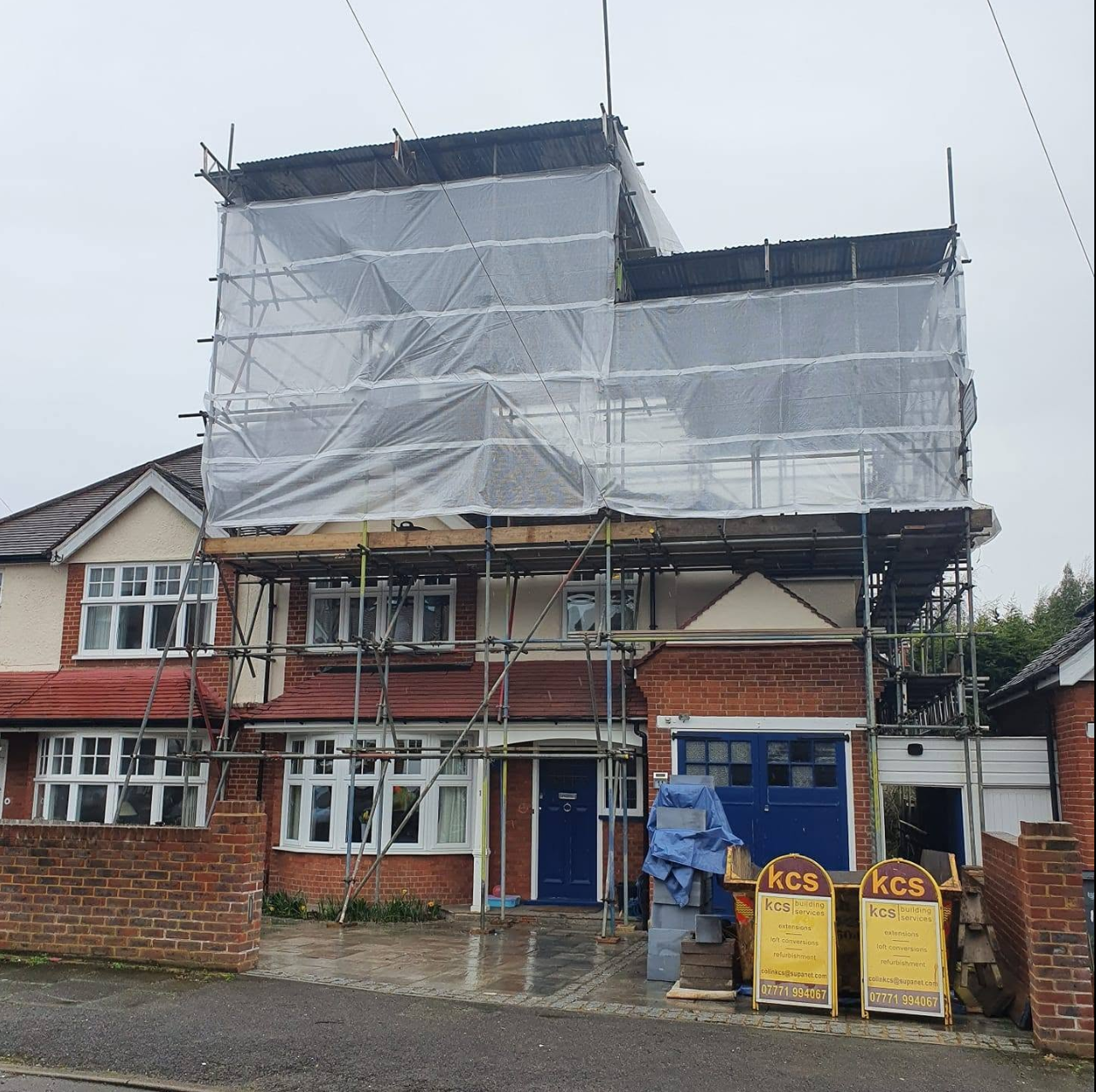 Semi-detached house with loft-height scaffold and clear shrink-wrapped temporary roof over a dormer loft conversion