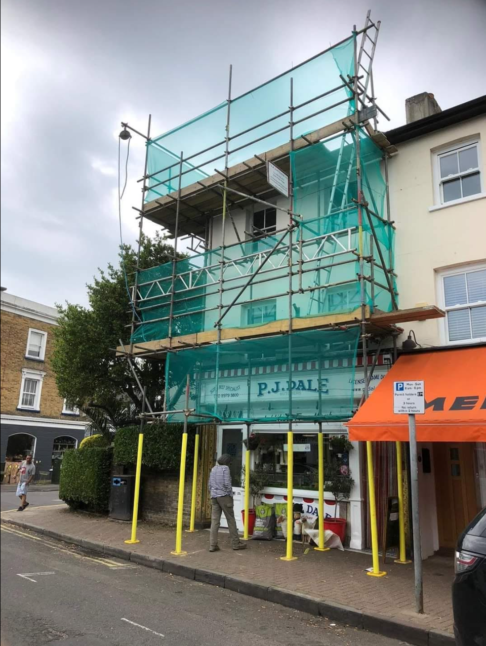 High-street shopfront with three-lift scaffold, green debris netting and yellow pedestrian standards